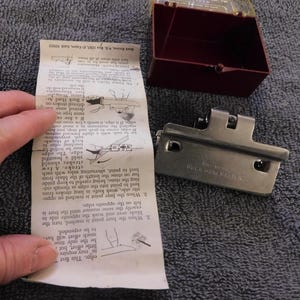 May include: A Buck HoneMaster knife sharpener with instructions. The sharpener is silver-colored and has a rectangular shape with a hinged design. A maroon plastic case and a folded instruction sheet are also present.