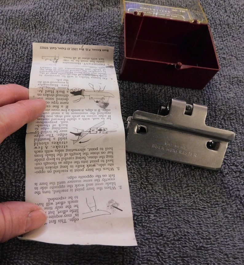 May include: A Buck HoneMaster knife sharpener with instructions. The sharpener is silver-colored and has a rectangular shape with a hinged design. A maroon plastic case and a folded instruction sheet are also present.