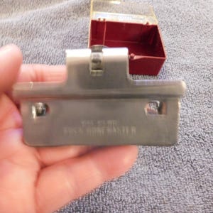 May include: A silver-colored metal cabinet hinge with the text "ROCK HOME MASTER" is held in a hand. A small, red plastic box with a clear lid is in the background. The hinge has two holes and a central mechanism.