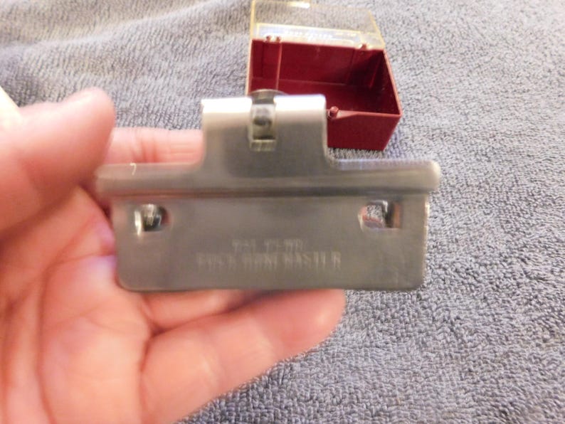 May include: A silver-colored metal cabinet hinge with the text "ROCK HOME MASTER" is held in a hand. A small, red plastic box with a clear lid is in the background. The hinge has two holes and a central mechanism.