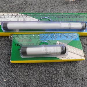 May include: Two clear plastic rolling rulers with black end caps, each featuring a central white cylinder with printed measurement markings. The rulers are positioned on their respective green and yellow cardboard packaging boxes.