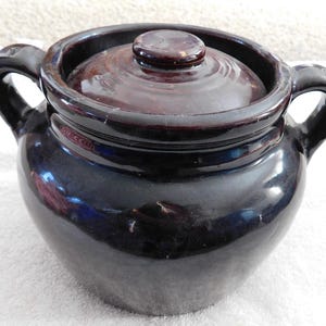 May include: A dark brown, glazed ceramic bean pot with a matching lid. The pot has two curved handles and a rounded body. The lid has a small, round knob. The pot's exterior has a glossy finish.