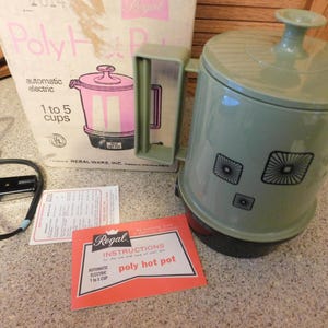 May include: A sage green Regal Poly Hot Pot with a matching lid and handle, featuring three square black graphic patterns on the side. The set includes a black power cord, a white cardboard box with pink text, and a red instruction manual for the 1 to 5 cup unit.