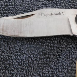K&ouml;nnte beinhalten: Nahaufnahme eines Klappmessers mit silberner Klinge und Holzgriff. Die Klinge ist mit "Nighthawk V" beschriftet. Das Messer ist teilweise ge&ouml;ffnet und zeigt die scharfe Kante. Ein kleiner, runder Silberniet ist am Griff sichtbar.