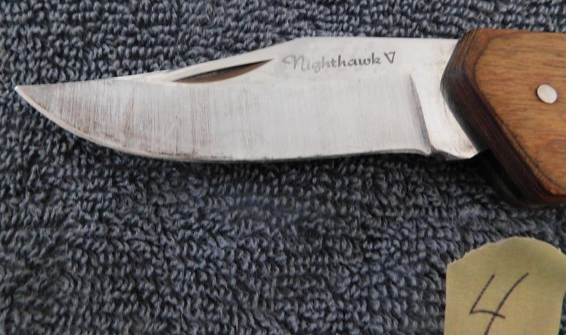 K&ouml;nnte beinhalten: Nahaufnahme eines Klappmessers mit silberner Klinge und Holzgriff. Die Klinge ist mit "Nighthawk V" beschriftet. Das Messer ist teilweise ge&ouml;ffnet und zeigt die scharfe Kante. Ein kleiner, runder Silberniet ist am Griff sichtbar.