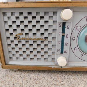 Puede incluir: Radio reloj Emerson vintage con marco dorado. La radio presenta una cubierta de altavoz a cuadros gris claro con el logotipo de Emerson en dorado. La esfera del reloj es azul claro con n&uacute;meros y manecillas negras. Dos perillas blancas est&aacute;n a la derecha.