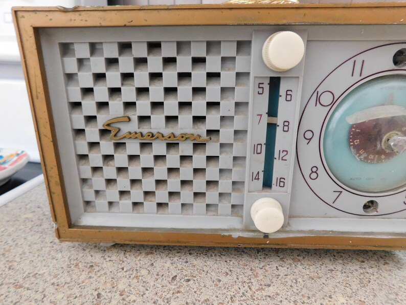 Puede incluir: Radio reloj Emerson vintage con marco dorado. La radio presenta una cubierta de altavoz a cuadros gris claro con el logotipo de Emerson en dorado. La esfera del reloj es azul claro con n&uacute;meros y manecillas negras. Dos perillas blancas est&aacute;n a la derecha.