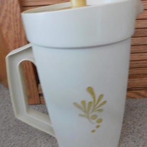 May include: A vintage white plastic pitcher with a handle and a lid. The pitcher has a yellow floral design on the side and a yellow top. The pitcher is designed to hold liquids.