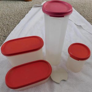 May include: A collection of clear plastic food storage containers with red lids. The set includes oval and cylindrical shapes, with a tall, slender container and a small round container. A white, circular lid is also present.