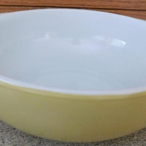May include: A vintage yellow and white mixing bowl with a rounded shape and two handles. The bowl has a smooth, glossy finish and is likely made of ceramic or glass. It is a classic kitchenware item.