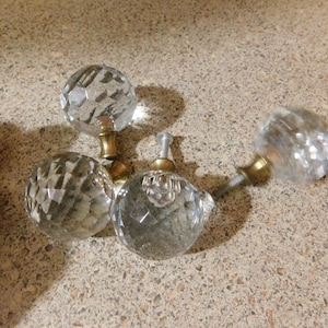 May include: Four clear crystal cabinet knobs with faceted surfaces and brass-colored bases. The knobs are arranged on a speckled, light brown surface. The knobs are likely for furniture or decorative use.