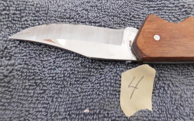 K&ouml;nnte beinhalten: Ein Edelstahlmesser mit Holzgriff. Die Klinge ist gebogen und hat eine scharfe Schneide. Der Griff ist braun und hat einen wei&szlig;en Punkt. Ein St&uuml;ck Klebeband mit der Nummer 4 befindet sich im Bild.