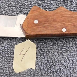 K&ouml;nnte beinhalten: Ein Klappmesser mit einer Klinge aus rostfreiem Stahl und einem Holzgriff. Der Griff ist mittelbraun und mit drei kleinen, wei&szlig;en Nieten befestigt. Ein kleines St&uuml;ck Papier mit der Zahl "4" befindet sich darunter.