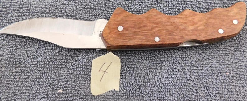 K&ouml;nnte beinhalten: Ein Klappmesser mit einer Klinge aus rostfreiem Stahl und einem Holzgriff. Der Griff ist mittelbraun und mit drei kleinen, wei&szlig;en Nieten befestigt. Ein kleines St&uuml;ck Papier mit der Zahl "4" befindet sich darunter.