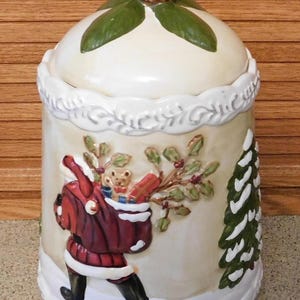 May include: A ceramic Christmas cookie jar with a lid topped with a pinecone and green leaves. The jar features a relief design of Santa Claus carrying a sack of toys and a Christmas tree.