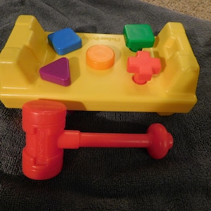 May include: A yellow plastic toy workbench featuring four colorful geometric shapes including a blue square, a purple triangle, an orange circle, and a green square, plus a red cross-shaped peg and a matching red plastic toy hammer.