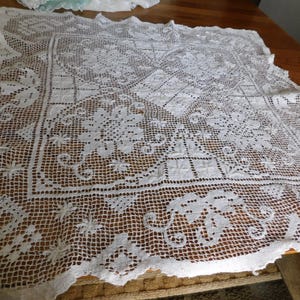May include: A white, open-weave lace tablecloth with a detailed floral and geometric pattern. The edges are scalloped, and the lace is laid out on a wooden surface. The tablecloth is likely vintage or handmade.