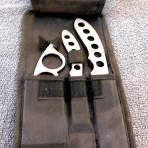 May include: A black nylon throwing knife set with three silver knives. The knives have a variety of shapes, including a ring, a small knife, and a larger knife with holes. The set is stored in a black nylon case.