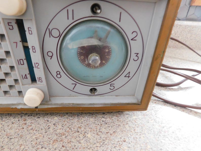Puede incluir: Primer plano de una radio vintage con reloj. La radio tiene una cara gris claro con un reloj con fondo azul claro y manecillas marrones. El marco de la radio es de color marr&oacute;n c&aacute;lido y tiene perillas blancas y un dial.
