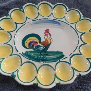 May include: A ceramic egg serving platter with a scalloped edge. The platter features a central rooster illustration with yellow, green, and blue accents. The outer rim has egg-shaped indentations, each painted yellow. The design is on a white background.