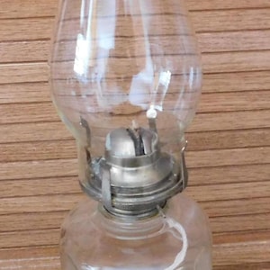 May include: A clear glass oil lamp with a metal burner assembly. The lamp has a rounded base, a bulbous midsection, and a cylindrical top. The lamp is designed to hold oil and a wick for illumination. The background is a wooden wall.