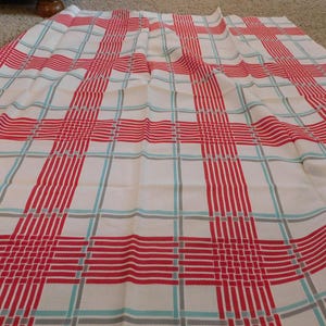May include: A rectangular fabric piece featuring a white background with a prominent plaid pattern. The design consists of bold red vertical and horizontal stripes, accented by thinner, intersecting light blue and grey lines throughout the textile.