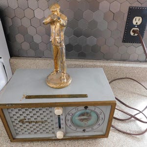 Puede incluir: Una radio Emerson vintage con una esfera de reloj y diales de sintonizaci&oacute;n, con una estatua dorada de un hombre. La radio tiene un cuerpo gris con un borde dorado y una rejilla de altavoz. La esfera del reloj tiene un dise&ntilde;o azul y blanco.