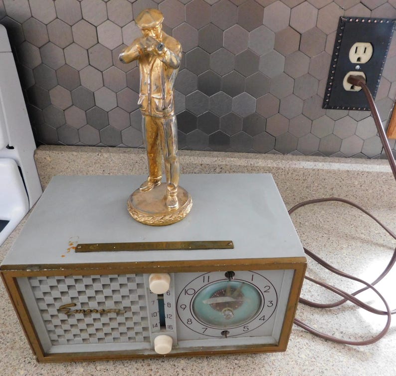 Puede incluir: Una radio Emerson vintage con una esfera de reloj y diales de sintonizaci&oacute;n, con una estatua dorada de un hombre. La radio tiene un cuerpo gris con un borde dorado y una rejilla de altavoz. La esfera del reloj tiene un dise&ntilde;o azul y blanco.