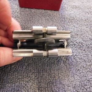 May include: A silver-toned metal object with a black rubber component, possibly a tool or clamp. The object has a screw mechanism and is held by a hand, with a red box visible in the background.