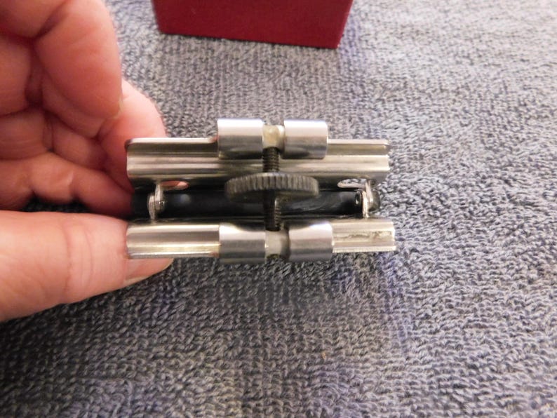 May include: A silver-toned metal object with a black rubber component, possibly a tool or clamp. The object has a screw mechanism and is held by a hand, with a red box visible in the background.