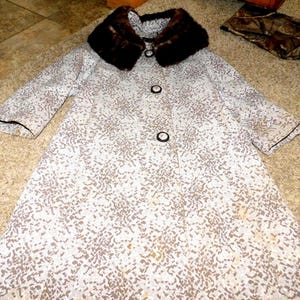 May include: A vintage coat with a speckled pattern in shades of white and brown. The coat features a dark brown fur collar, three black buttons, and black trim on the sleeves. The coat is a knee-length style.