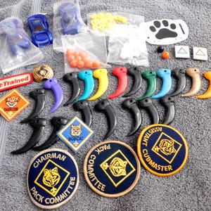 May include: A collection of scouting items including three blue toy cars in plastic bags, orange and white beads, a white paw print cutout, two small square tiles, embroidered patches, and numerous colorful plastic claw-shaped pendants.