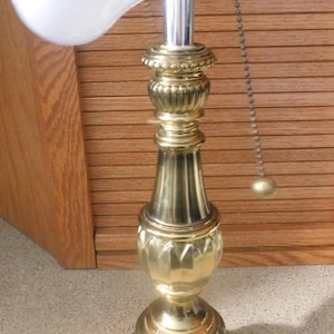 May include: A brass table lamp with a polished metallic finish, featuring a tiered, decorative base and a central column with ornate detailing. The lamp includes a white light bulb, a brass socket, and a pull chain with a small spherical end.