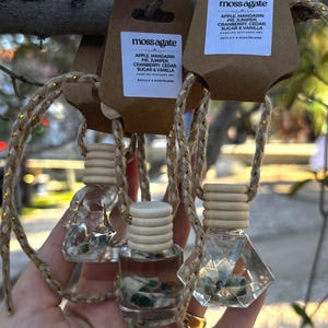 Puede incluir: Tres botellas difusoras colgantes con el texto "moss agate" y descripciones de fragancias. Las botellas son de vidrio transparente con tapas de madera y están suspendidas por una cuerda dorada.