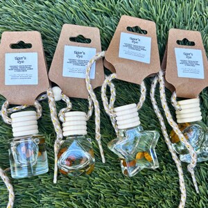 May include: Four clear glass car air fresheners with wooden tops and hanging cords. Each has a different shape, including a star and a square. The tags read "tiger's eye" and list ingredients. The fresheners are on a bed of green grass.