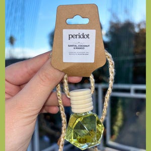 Könnte beinhalten: Ein hängender Auto-Lufterfrischer mit einer geometrischen Glasflasche, die eine gelbe Flüssigkeit enthält. Die Flasche wird von einem beigefarbenen Seil und einem braunen Etikett mit dem Aufdruck "peridot" und "Santal Coconut & Mango" gehalten.