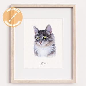 May include: A framed watercolor painting of a cat with gray and white fur, green eyes, and a pink nose. The artwork is signed "Clee" in cursive. The frame is light-colored wood, and the artwork is labeled "Hand-Painted Original Artwork".