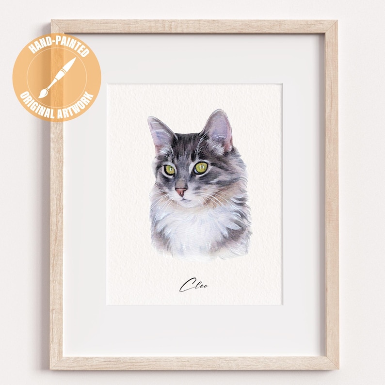 May include: A framed watercolor painting of a cat with gray and white fur, green eyes, and a pink nose. The artwork is signed "Clee" in cursive. The frame is light-colored wood, and the artwork is labeled "Hand-Painted Original Artwork".