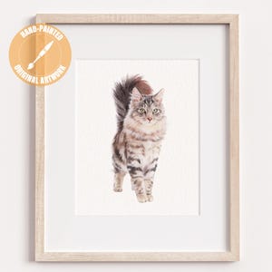 May include: A framed watercolor painting of a fluffy cat with gray and brown stripes, standing on a white background. The cat has a long, bushy tail and is facing forward. The frame is light-colored, and the artwork is labeled "Hand-Painted Original Artwork."