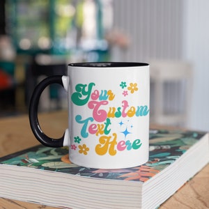 May include: White ceramic mug with a black handle and black rim. The mug has a colorful retro design with the text "Your Custom Text Here" and small flower and star accents.