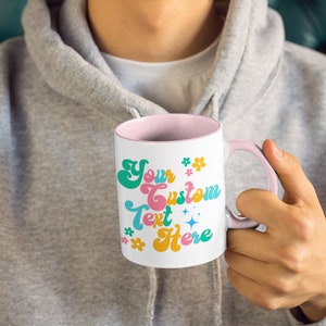 May include: White ceramic mug with a pink rim and handle. The mug has a retro-style design with the text "Your Custom Text Here" in colorful letters and flowers.