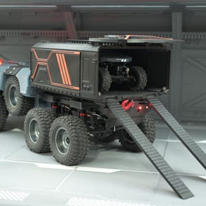 May include: A futuristic, multi-wheeled transport vehicle with a black cargo area and orange accents. A smaller all-terrain vehicle is inside. The transport has a ramp for loading and unloading. The vehicle is grey and black.