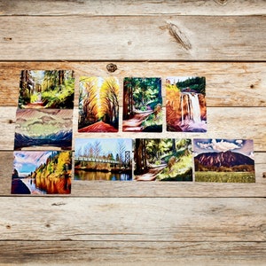 May include: A collection of nine landscape photographs featuring various scenes of nature, including a forest path, a bridge over a river, a waterfall, and a mountain range. The photos are printed on a textured paper and have a vintage aesthetic.