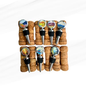 May include: A set of eight silver bottle stoppers with colorful images of landscapes and cityscapes. The stoppers are displayed on top of cork wine bottle stoppers.