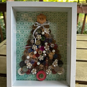 Puede incluir: Una obra de arte enmarcada de un árbol de Navidad hecha con varios botones coloridos. El árbol está dispuesto sobre un fondo estampado y adornado con elementos decorativos. El marco blanco añade un borde limpio a la obra, adecuado para la decoración navideña.