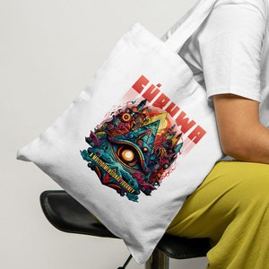 Puede incluir: Bolso tote blanco con un diseño gráfico colorido, incluyendo la palabra "Ehdwa" en rojo. El bolso lo sostiene una persona que lleva una camiseta blanca y pantalones amarillos. El diseño presenta un ojo y formas abstractas.
