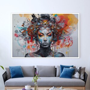 May include: A colorful abstract painting of a woman's face with a floral crown. The painting is framed in white and hangs on a white wall above a gray sofa with blue pillows.