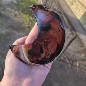 May include: A polished crescent moon shaped stone carving featuring a swirling pattern of deep mahogany, dark brown, and cream coloured bands. The surface is smooth and reflective, showcasing natural mineral layers with a glossy finish.