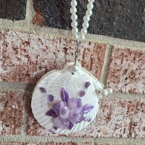 May include: A white seashell pendant adorned with purple amethyst crystals and a pearl necklace. The seashell is decorated with a cluster of amethyst crystals in the center and smaller crystals around the edges. The necklace is made of white pearls.