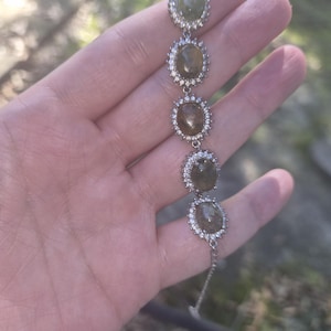 May include: A silver-toned bracelet featuring five oval-shaped, translucent olive-green cabochon stones. Each stone is surrounded by a halo of small, clear, sparkling round-cut stones set in a metallic frame, connected by thin silver-toned links.